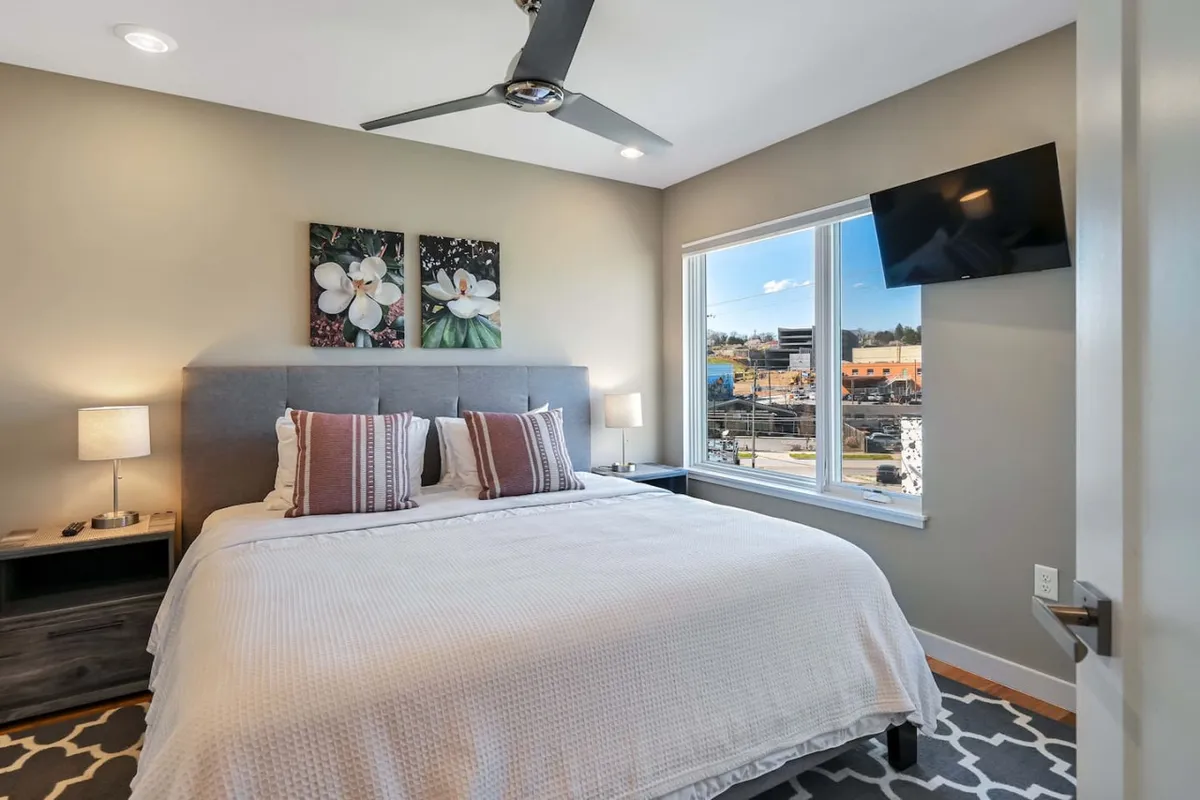 A tranquil bedroom space that includes a comfortable bed, soft wall art, a window allowing daylight to pour in, nightstands for convenience, and a wall-mounted TV for entertainment. The room exudes a calm, relaxing atmosphere ideal for unwinding.