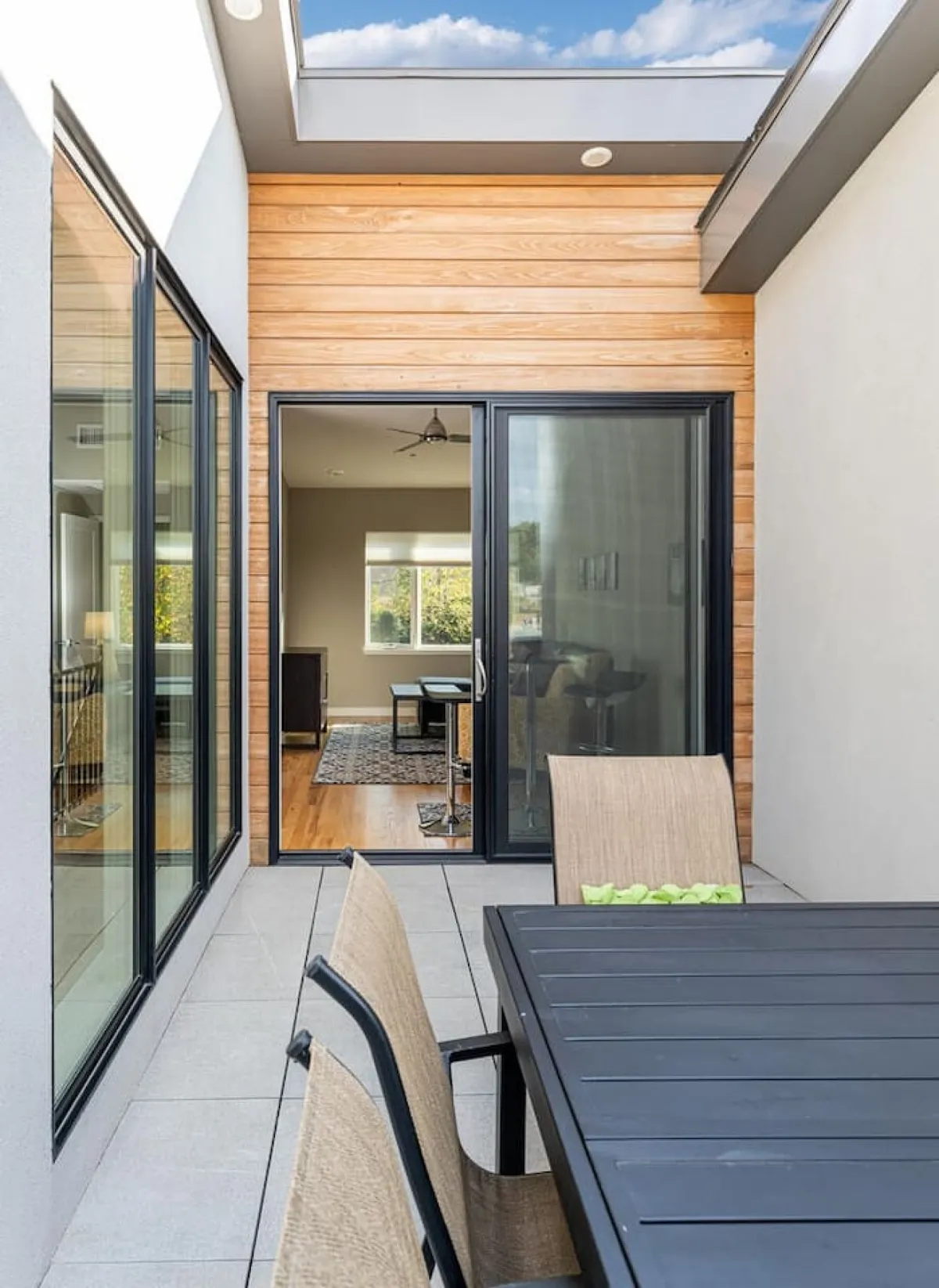 This image showcases a dining table set next to a sliding glass door that opens out to a patio or balcony with outdoor chairs. The scene blends indoor and outdoor environments, inviting guests to enjoy al fresco dining or relaxing with a book. The wooden wall adds warmth, while the outdoor setup suggests possibilities for rooftop or patio retreats.