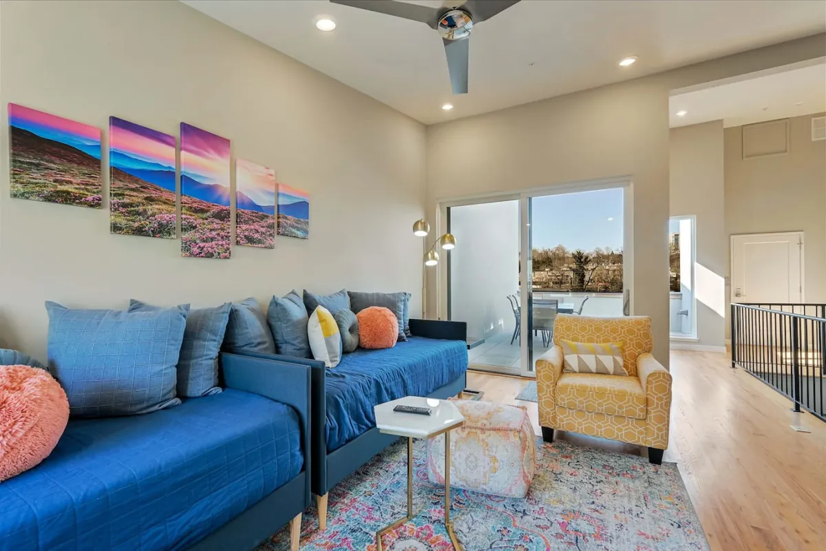 Featuring a blue sofa, colorful artwork, a yellow armchair, and a coffee table positioned by a sliding glass door, this scene highlights a cheerful living area with direct access to the outdoors. The sliding glass door indicates the ease of moving between cozy indoor spaces and the exterior environment, reinforcing the multi-story, multi-functional design of the home.