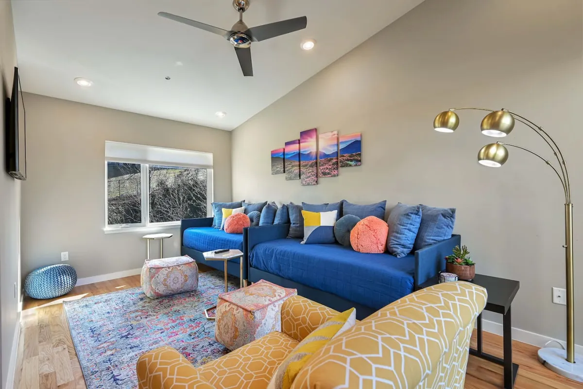 A bright and cheerful living space with a large blue sectional sofa, colorful throw pillows, decorative artwork, and comfortable poufs. The layout invites relaxation and social interaction, ideal for groups returning from city adventures.