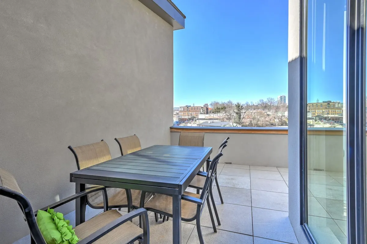 An exterior rooftop patio setup featuring a dining table and chairs arranged to take in expansive city views, with the horizon stretching towards partial mountain silhouettes. The mood is inviting and casual, perfect for both relaxing and social gatherings as sunset light bathes the space.