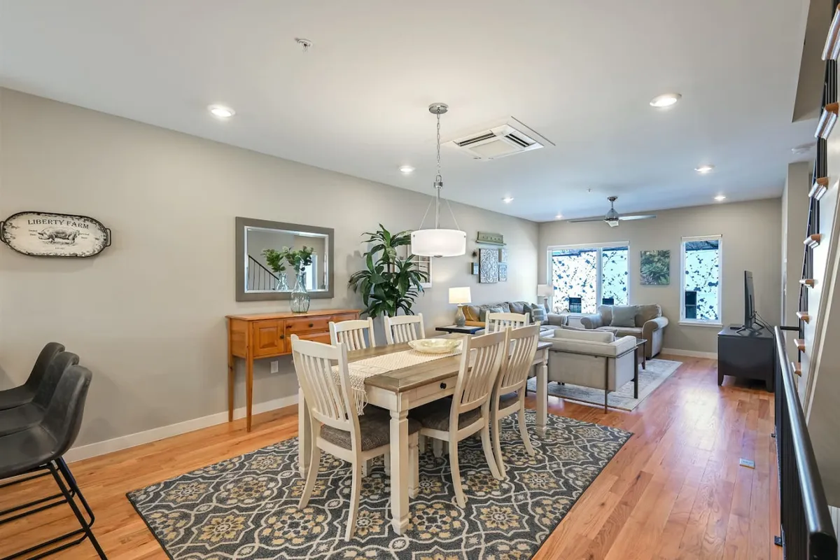 A spacious open-concept area featuring a large dining table set for a group meal, with a living room seating area visible in the background and decorative plants adding warmth. The arrangement encourages gathering and shared experiences.