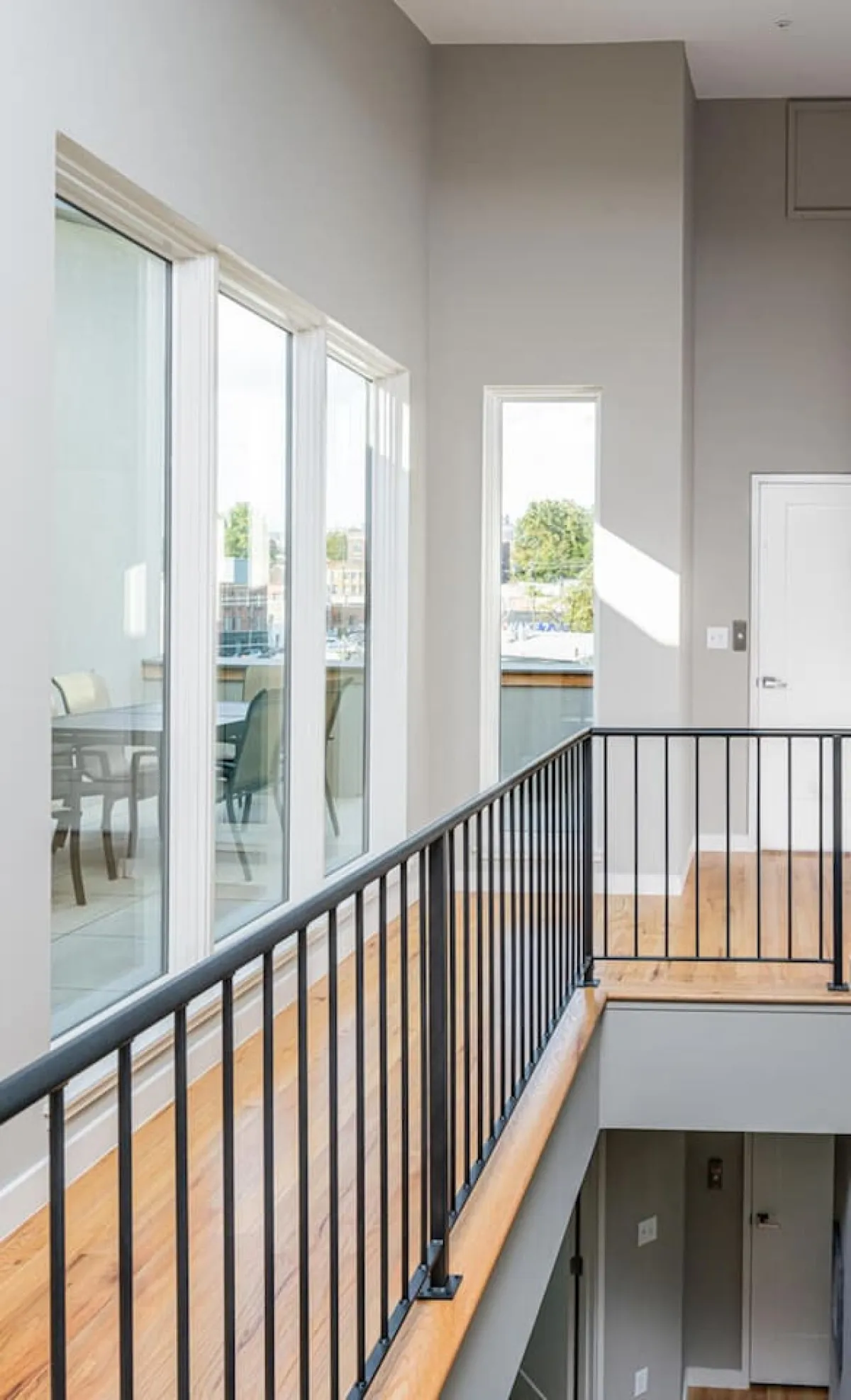 This image captures a bright, open space with large windows that flood the room with natural light. The wooden railing hints at a multi-level layout, and the open concept design offers views beyond the immediate interior. The scene feels airy and welcoming, suggesting a seamless flow between indoor comfort and outdoor vistas. The suggestion of a balcony visible through the windows subtly connects the interior to the outside world.