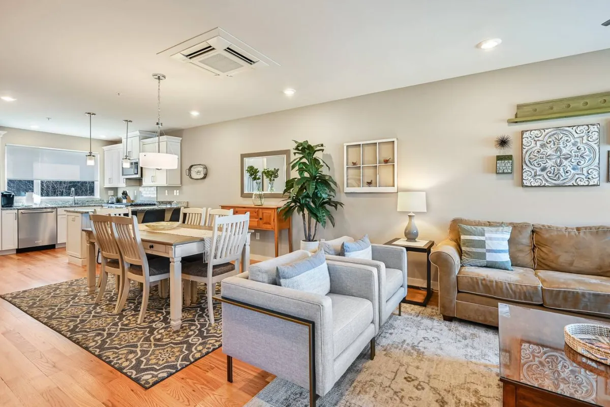 This image presents a warmly lit interior scene encompassing a dining table and a comfortable sofa, with a green plant and decorative wall art enhancing the inviting atmosphere. The arrangement suggests a space ideal for both family dinners and relaxed social gatherings, with thoughtful decor that makes the area feel both lively and intimate.